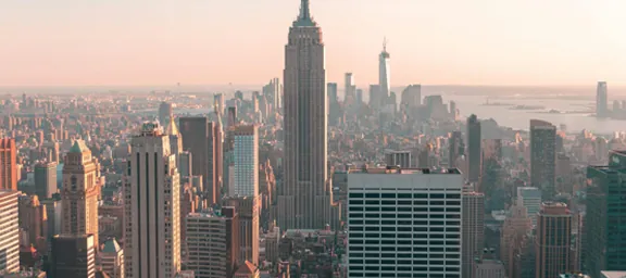 A high-angle, wide view of the New York City skyline during the golden hour. The Empire State Building stands prominently in the center, surrounded by numerous skyscrapers, all bathed in a warm, pinkish-orange sunset glow with the Hudson River visible in the distance.