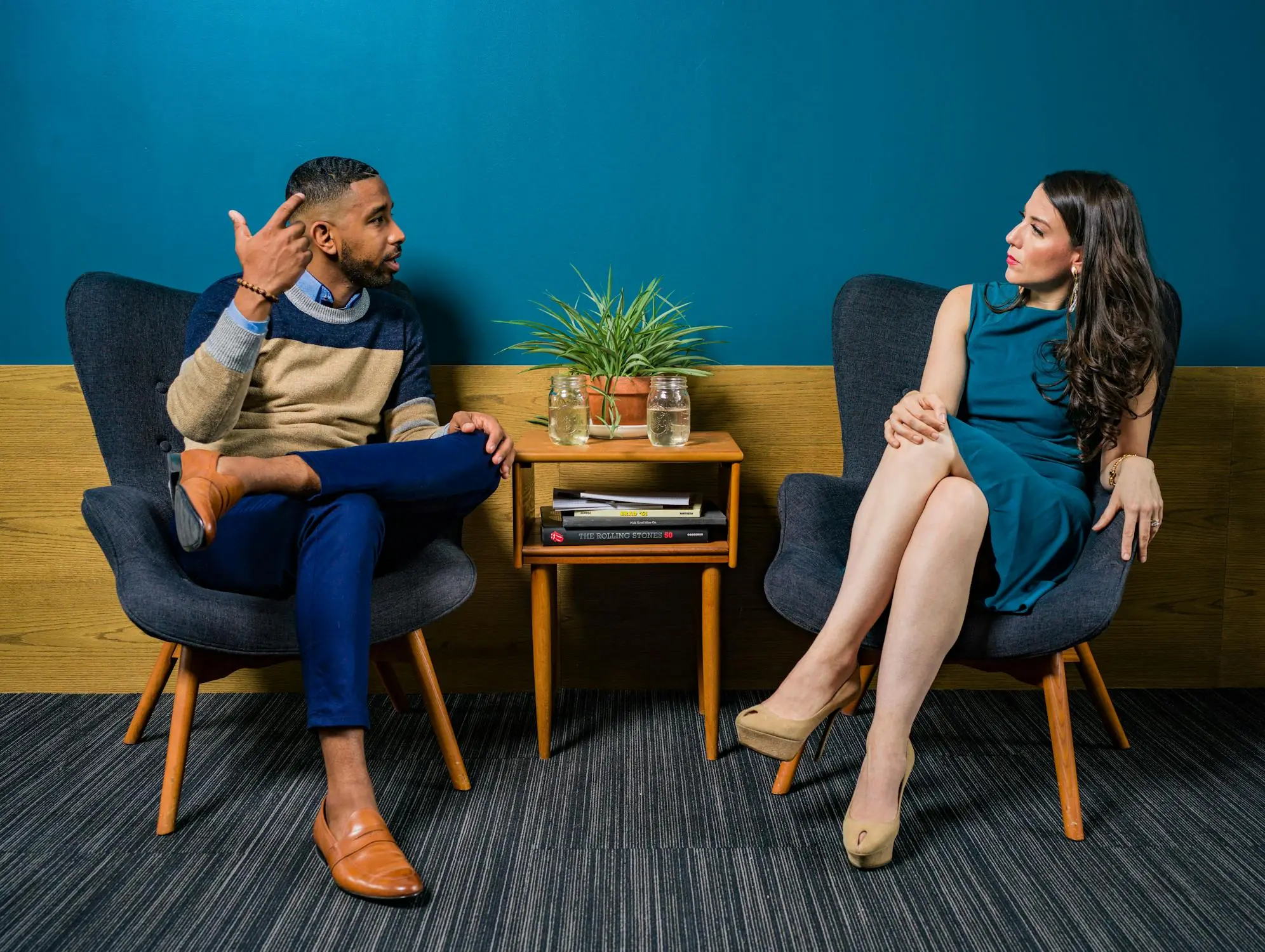 A man and a woman engaged in a professional discussion or interview, seated in modern grey armchairs. They are positioned on either side of a small wooden end table with a potted plant and books. The background features a striking teal wall and light wood paneling, creating a clean and contemporary office atmosphere.